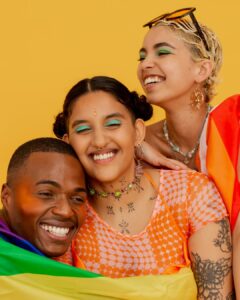 Three joyful people with brightly colored makeup and accessories smile and embrace in front of a yellow background. They are wrapped in Pride flags, expressing warmth, celebration, and togetherness.