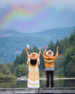 Two Indigenous Canadian people singing and dancing by a lake with a rainbow in the distance. One holds a musical drum, they both have their hands outstretched and raised in the air.