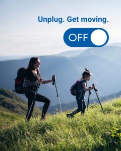 A woman and young girl hike through a field, both equipped with poles and backpacks. Mountains are seen in the background. The image says “Unplug. Get moving.”