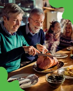 Holiday gathering featuring two older adults carving a ham around a dining room table smiling and enjoying festive celebrations.